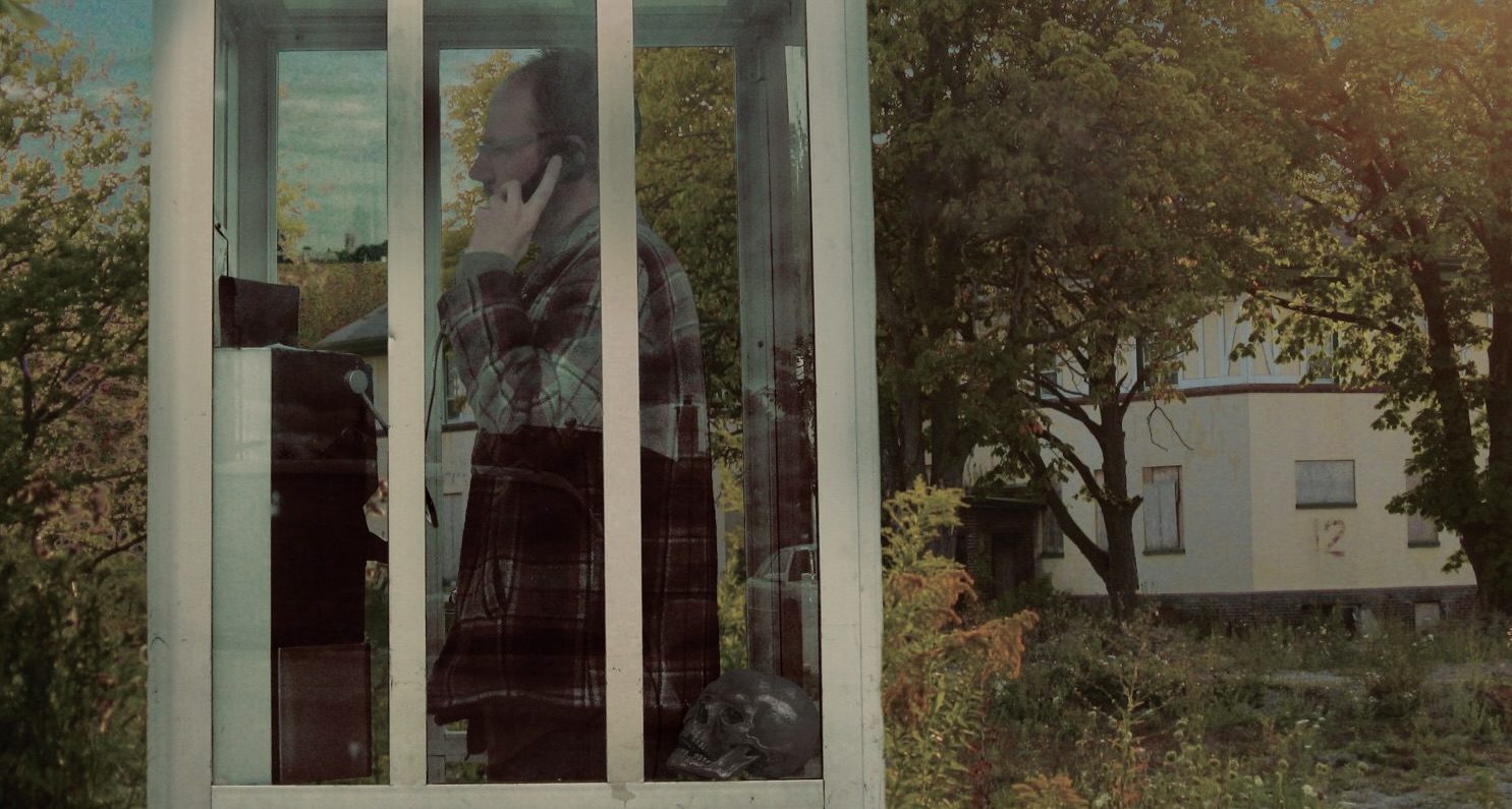 Black Schwartz standing in a glass phone booth with a skull resting nearby &ndash; promotional photo for Little Meadow